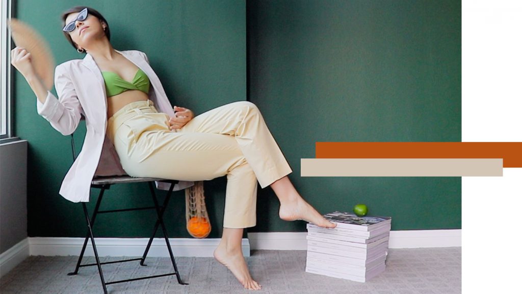 Woman wearing yellow trousers, green bikini top, pink blazer while looking at the window